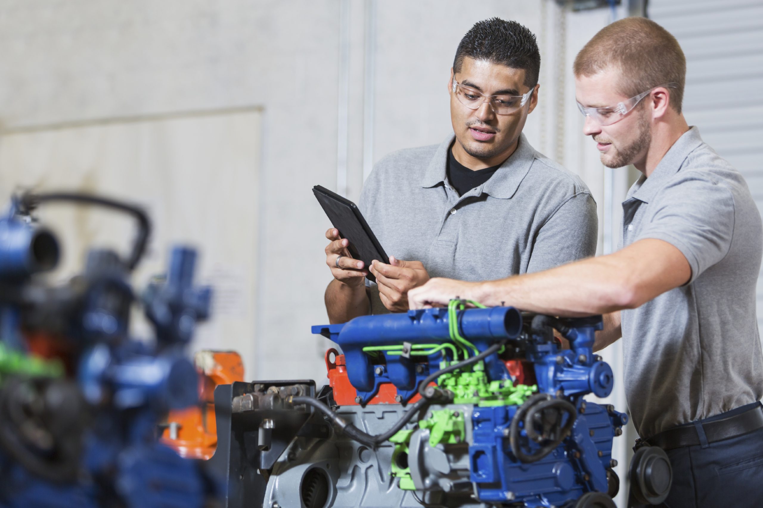 Two multi-ethnic young men in vocational school, taking a class on reparing diesel engines.  They are working on an engine that has had parts painted different colors for training purposes.  They are wearing safety glasses. The Hispanic man is reading a digital tablet.